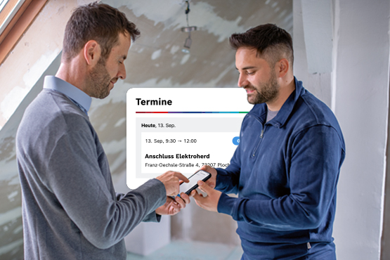 Zwei Männer in hellen Räumen, einer zeigt auf ein Handy, im Hintergrund eine Wand mit Trockenbau.