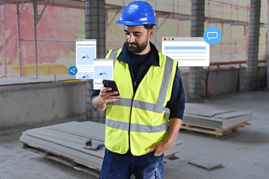 Ein Bauarbeiter in gelber Warnweste und blauer Schutzhelm schaut auf sein Smartphone, umgeben von Platten.