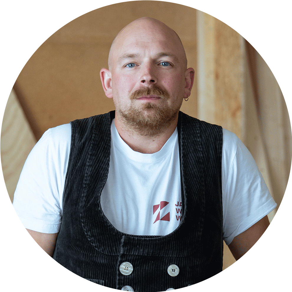 Ein Mann mit Glatze und Bart trägt ein schwarzes Weste über einem weißen T-Shirt. Holz im Hintergrund.