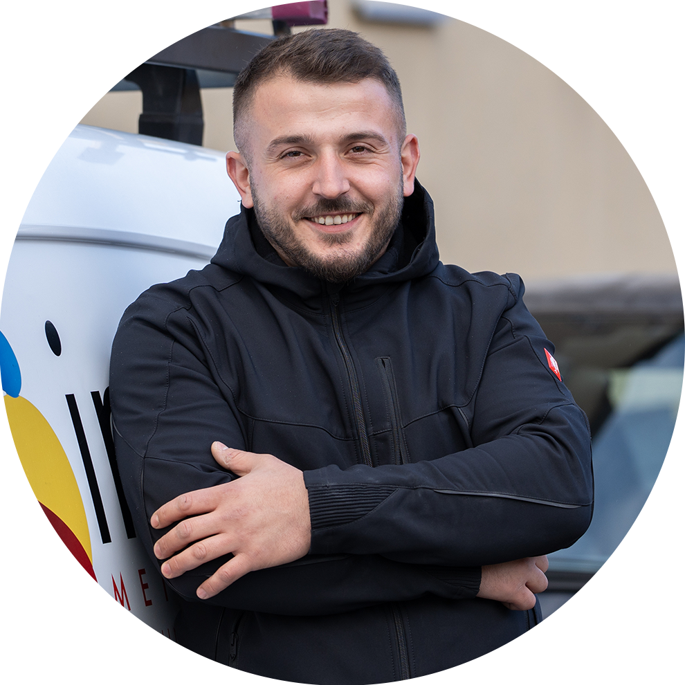 Ein lächelnder Mann in schwarzer Jacke steht vor einem weißen Lieferwagen mit buntem Logo. Er hat die Arme verschränkt und wirkt freundlich.