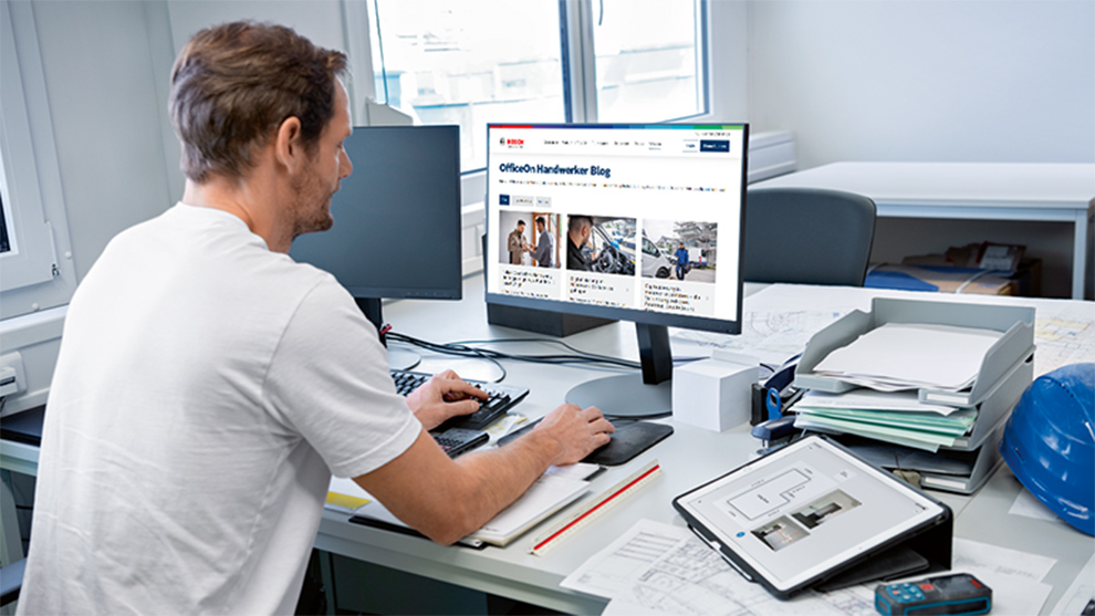 Ein Mann arbeitet am Computer in einem Büro mit Plänen und Werkzeug.