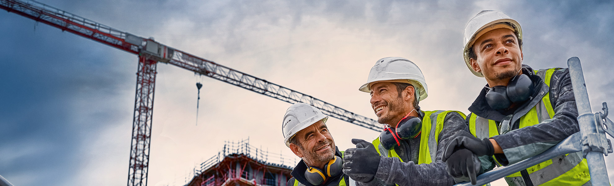 Drei Bauarbeiter mit Helmen und Warnwesten lächeln vor einem Kran.